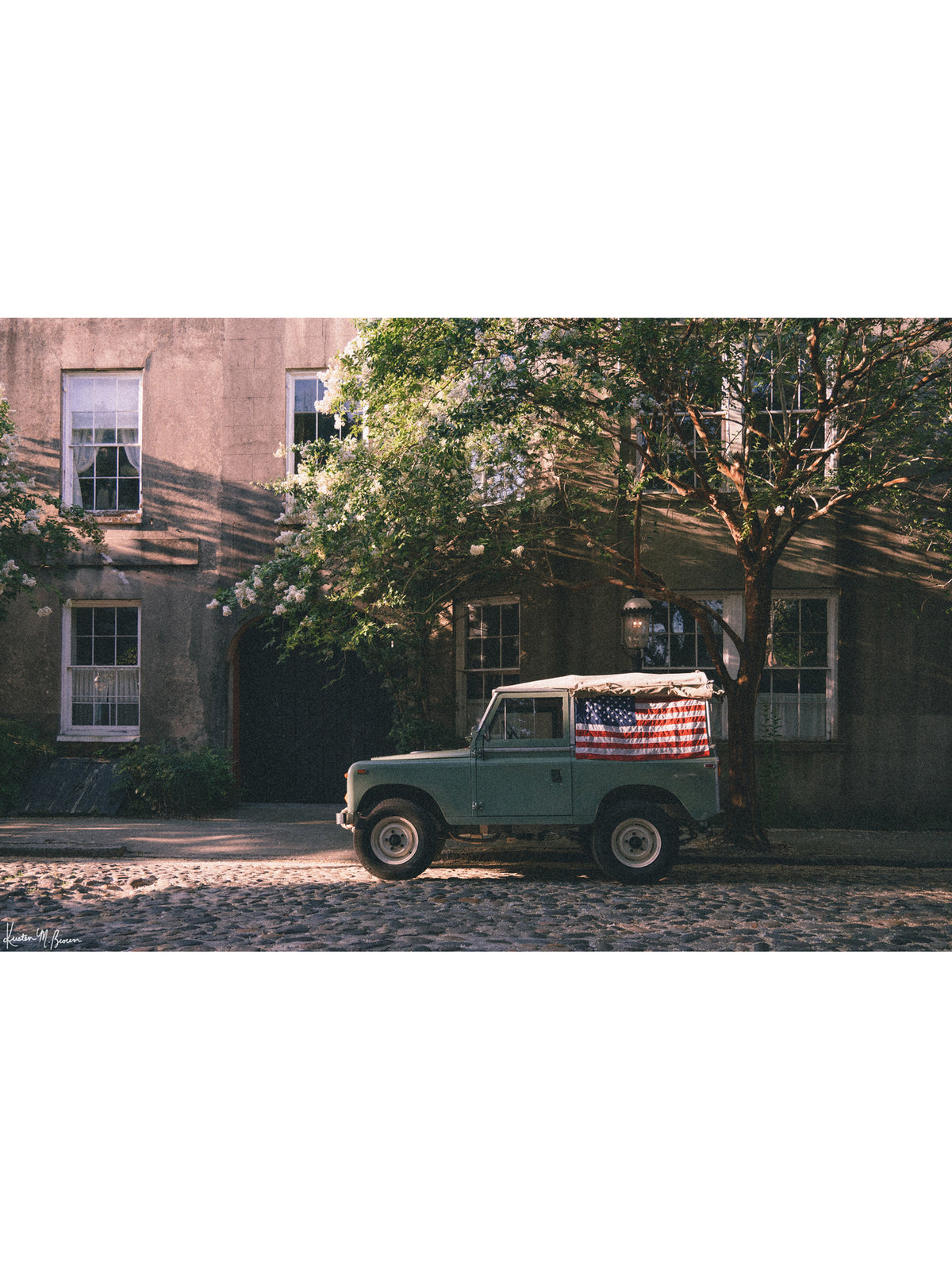 A vintage green Land Rover Series with an American flag draped over the rear window, parked on a cobblestone street in historic Charleston with blooming white Crepe Myrtle flowers. "Stars & Stripes & Series" is a journey into the charming streets of historic Charleston, where beauty and nostalgia meet. Vintage Land Rover photo print by Kristen M. Brown of Samba to the Sea for The Sunset Shop.