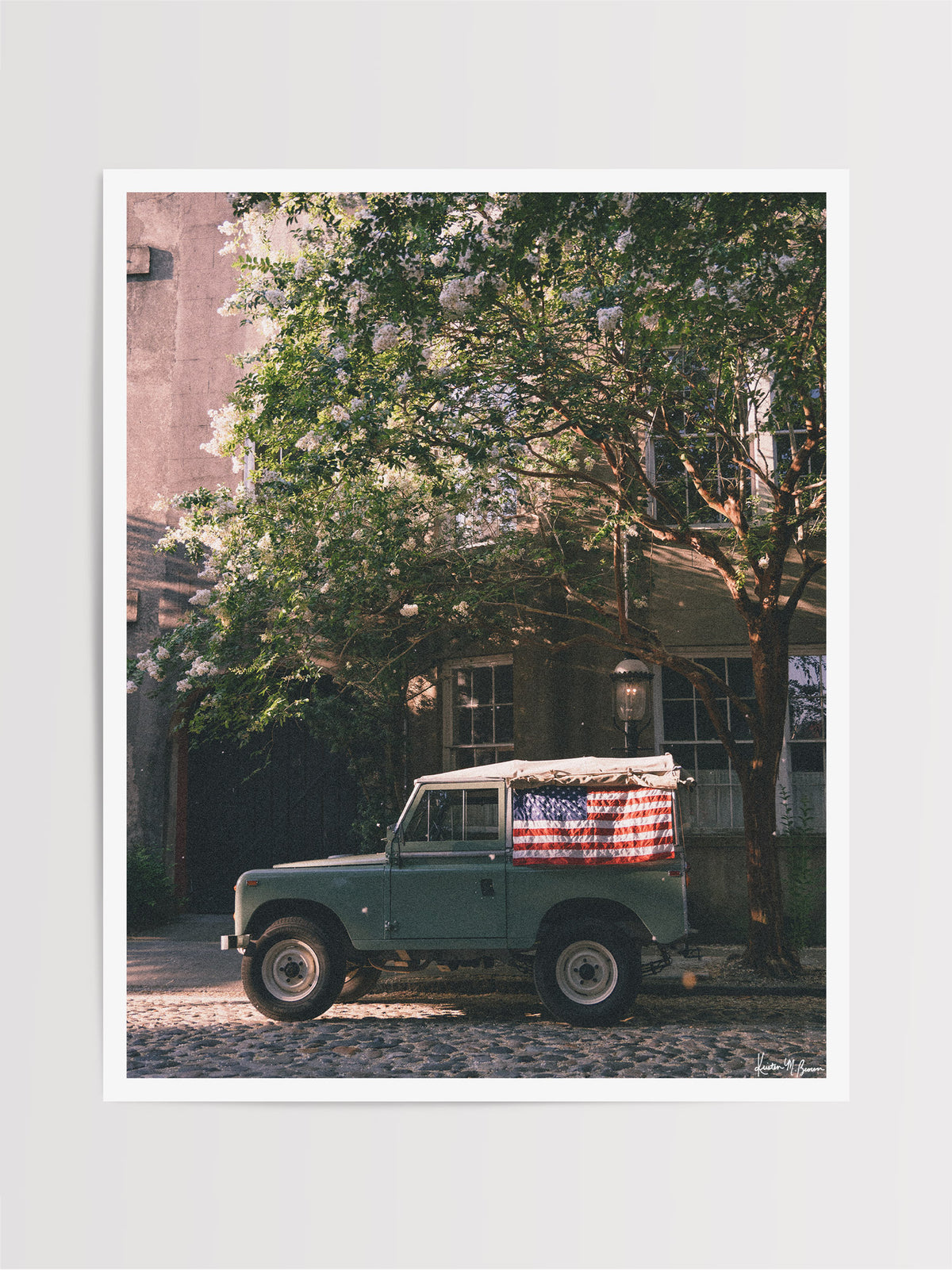 A vintage green Land Rover Series with an American flag draped over the rear window, parked on a cobblestone street in historic Charleston with blooming white Crepe Myrtle flowers. "Stars & Stripes & Series" is a journey into the charming streets of historic Charleston, where beauty and nostalgia meet. Vintage Land Rover photo print by Kristen M. Brown of Samba to the Sea for The Sunset Shop.