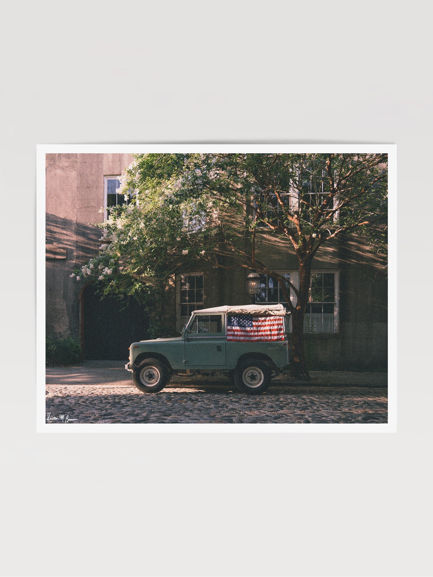 A vintage green Land Rover Series with an American flag draped over the rear window, parked on a cobblestone street in historic Charleston with blooming white Crepe Myrtle flowers. "Stars & Stripes & Series" is a journey into the charming streets of historic Charleston, where beauty and nostalgia meet. Vintage Land Rover photo print by Kristen M. Brown of Samba to the Sea for The Sunset Shop.