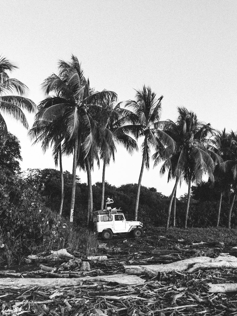 "Surf Bandida" black and white photo print of surfer girl checking the surf with her vintage Toyota FJ40 Land Cruiser racked up with surfboards among the palm trees in Costa Rica. Photographed by Costa Rica photographer Kristen M. Brown of Samba to the Sea for The Sunset Shop.