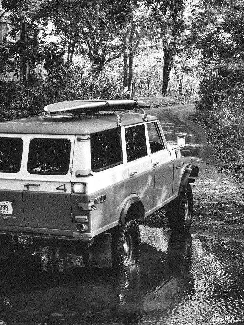 Over a river and down a dirt back road dirt lies a dreamy, peeling wave that only you and your surf amigos know about. You hear a new swell is on the way, so you message your friends to meet you at your secret spot, hop in your vintage Land Cruiser series 55, and surf the day away with just your surf amigos. Welcome back to your surf dream, all from the comfort of your home...wherever home may be with black and white photo print “Surf Cruiser&quot;. By Kristen M. Brown of Samba to the Sea @ The Sunset Shop.