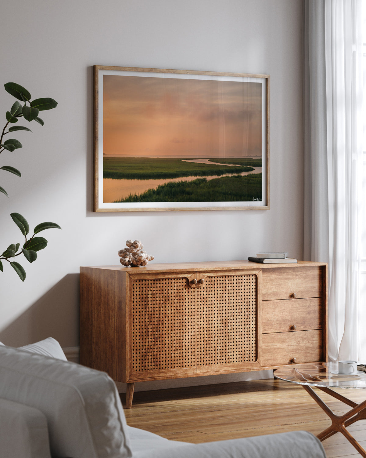 A scenic landscape photograph of a winding creek flowing through the marsh during sunrise, with the soft peach and pink clouds in the sky above and reflecting in the waters. "Tidal Glow" fine art photography print photographed by Kristen M. Brown, Samba to the Sea. Famed landscape art print styled on a light gray wall above a mid-century modern wooden credenza in a living room setting.