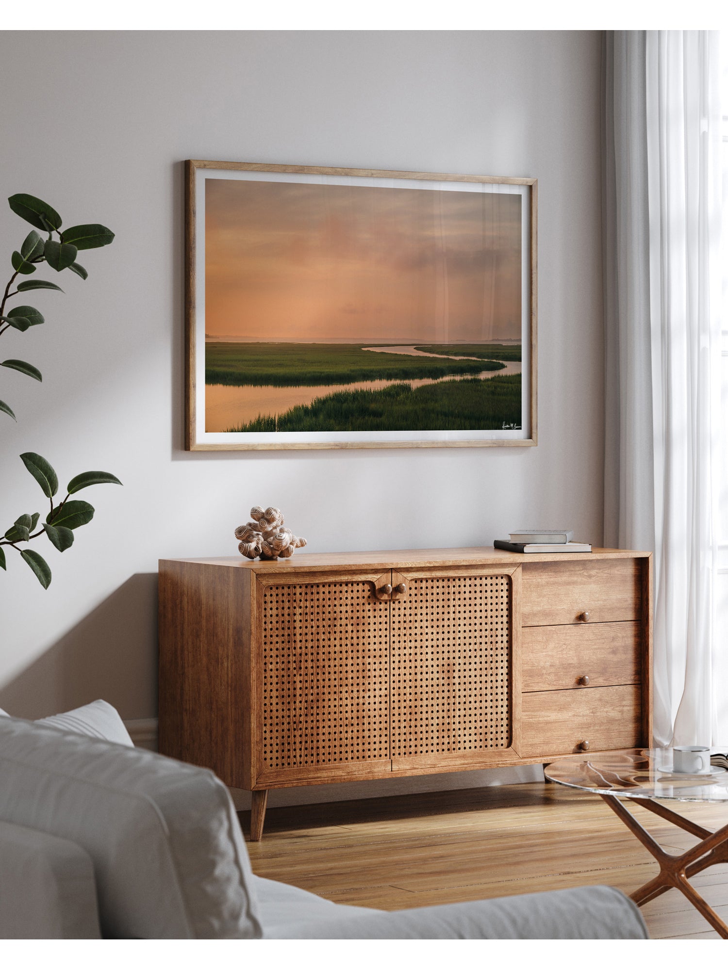 A scenic landscape photograph of a winding creek flowing through the marsh during sunrise, with the soft peach and pink clouds in the sky above and reflecting in the waters. "Tidal Glow" fine art photography print photographed by Kristen M. Brown, Samba to the Sea. Famed landscape art print styled on a light gray wall above a mid-century modern wooden credenza in a living room setting.
