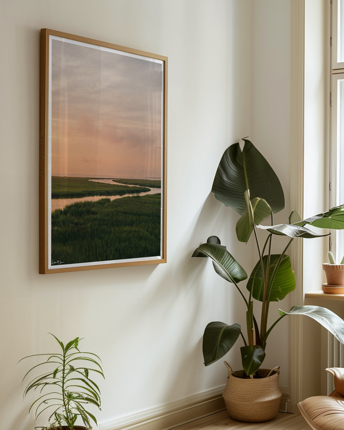 A scenic landscape photograph of a winding creek flowing through the marsh during sunrise, with the soft peach and pink clouds in the sky above and reflecting in the waters. "Tidal Glow" fine art photography print photographed by Kristen M. Brown, Samba to the Sea. Framed landscape art print styled on a cream wall in a bright room next to potted green plants.