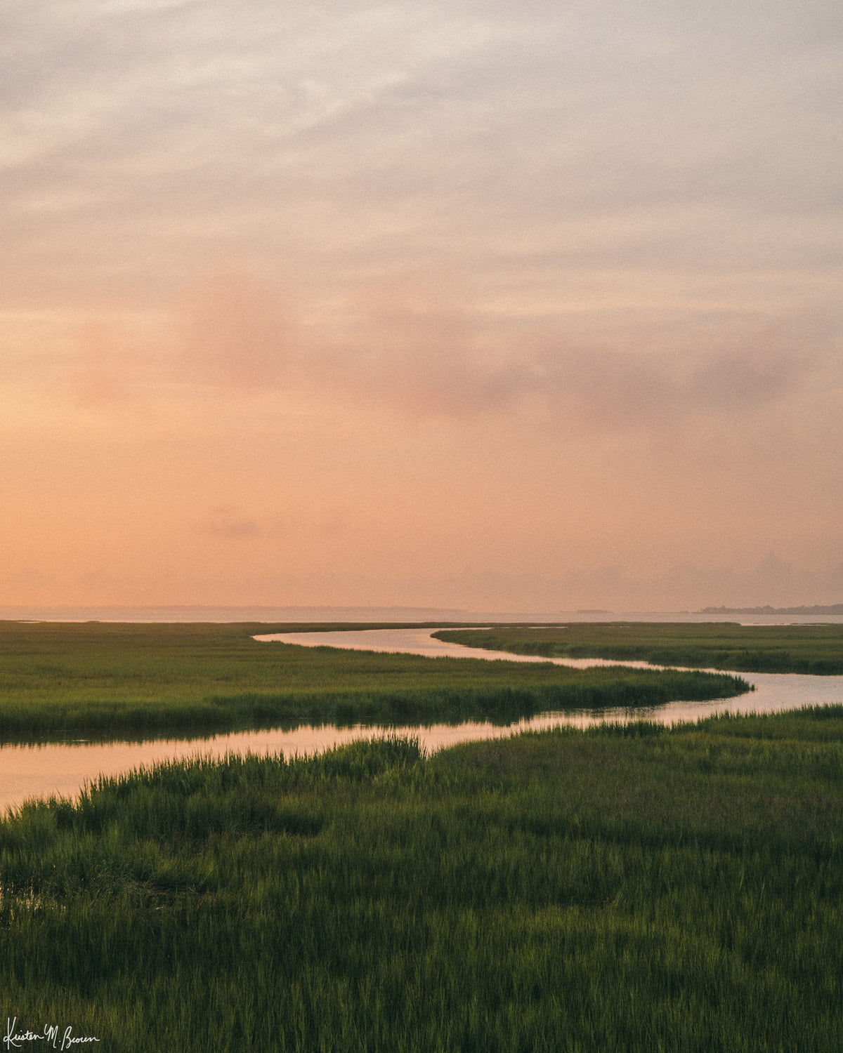 A scenic landscape photograph of a winding creek flowing through the marsh during sunrise, with the soft peach and pink clouds in the sky above and reflecting in the waters. "Tidal Glow" fine art photography print photographed by Kristen M. Brown, Samba to the Sea.