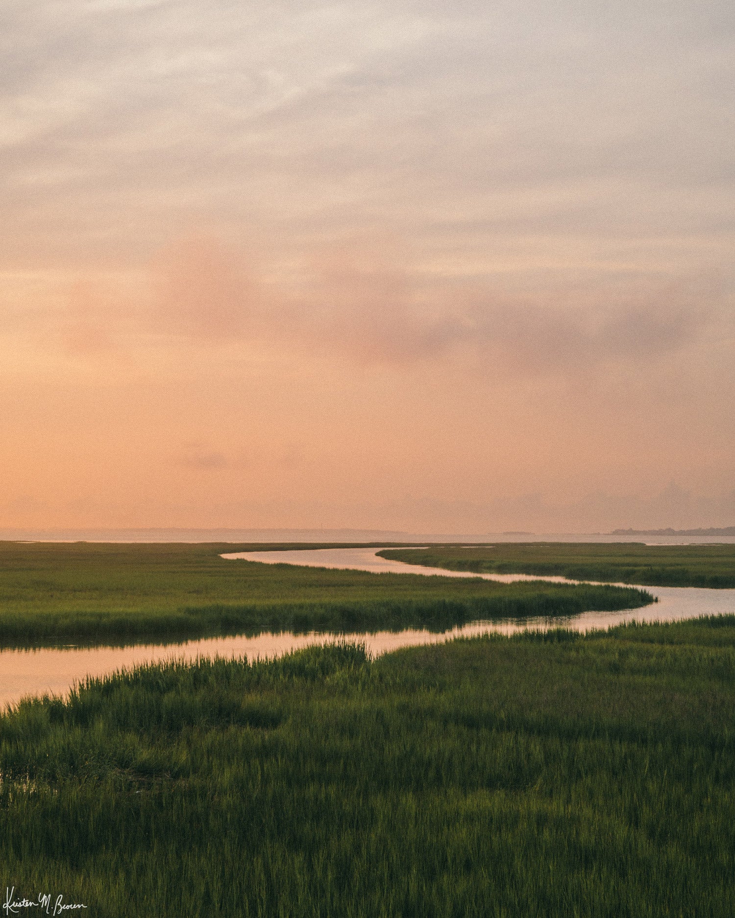 A scenic landscape photograph of a winding creek flowing through the marsh during sunrise, with the soft peach and pink clouds in the sky above and reflecting in the waters. "Tidal Glow" fine art photography print photographed by Kristen M. Brown, Samba to the Sea.