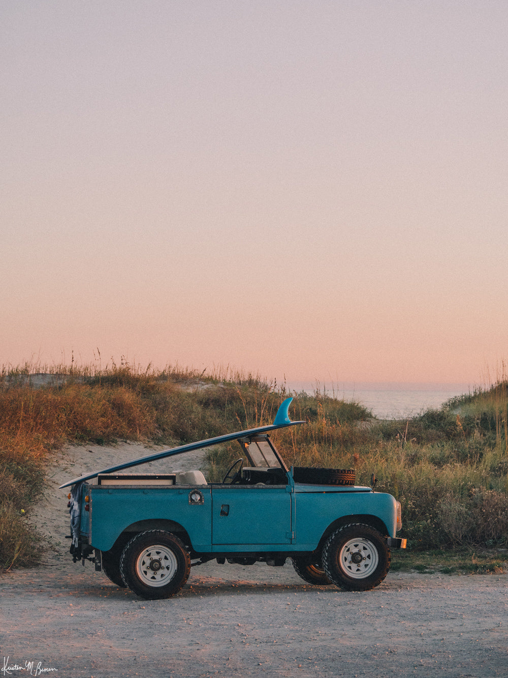 Photograph of vintage Land Rover series truck parked at the beach with a classic single fin surfboard amidst a pastel pink sunset sky. "Wave Chaser" photographed by Kristen M. Brown, Samba to the Sea.