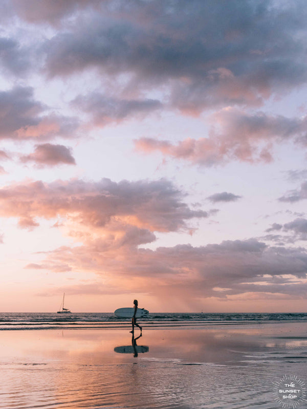 Angel del Mar - Sea Angel sunset surfer girl print by Samba to the Sea at The Sunset Shop. Photo of a female surfer walking on the beach during a pastel pink sunset in Tamarindo, Costa Rica.