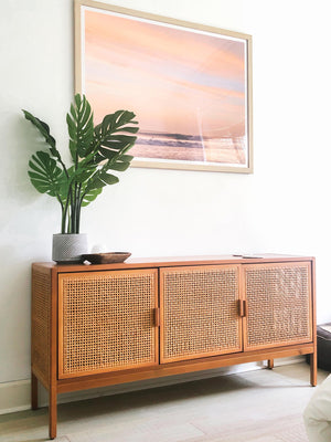 Pastel sunset wave framed photo print. Rattan console and Monstera plant for all the tropical boho vibes. "Hola Ola! - Hello Wave" Costa Rica sunset wave photo print photographed by Kristen M. Brown of Samba to the Sea for The Sunset Shop.