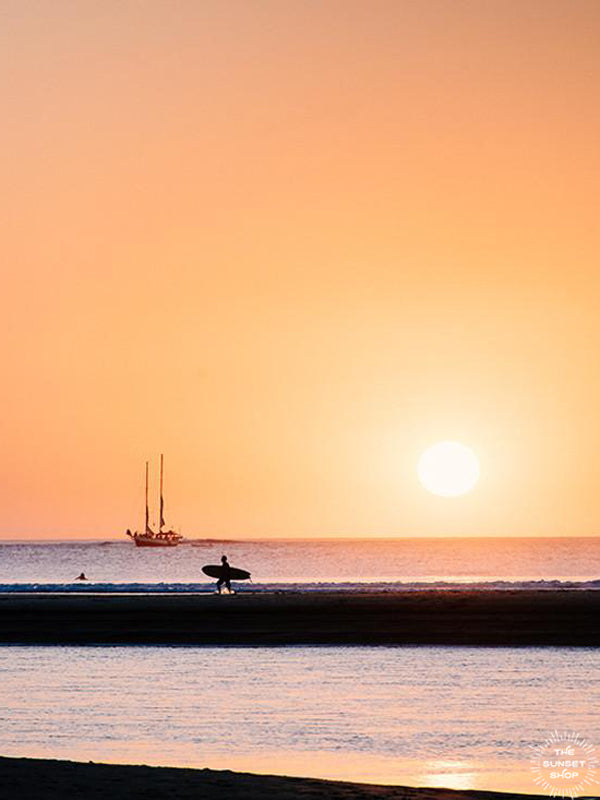 Magic Moments surfer during sunset print by Samba to the Sea at The Sunset Shop. Photograph of a surfer walking on the beach with a schooner sailboat sailing during sunset in Tamarindo, Costa Rica. 