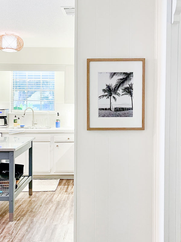 Coastal living kitchen with palm trees and surfboard black and white photo print. Transport yourself to your surf paradise with this black and white image of your surfboards - your magic sticks - patiently waiting under palm trees for you and your surf amigo/a to paddle out! Black and white image of two Robert August surfboards waiting to paddle out while laying under palm trees in Costa Rica. "Magic Sticks" palm tree surf print photographed by Kristen M. Brown of Samba to the Sea for The Sunset Shop.