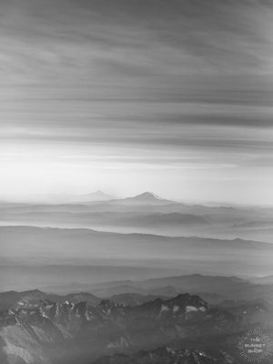 Black and white aerial image of late afternoon sun rays over Mt Rainier in Washington. What a glorious farewell the late afternoon sun gives the mountains. Magical late afternoon sun rays falling over Mt Rainier and the Cascade mountain range in Washington.  