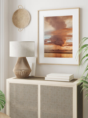 Gorgeous sunset photo print hanging above cane credenza in a tropical modern room. “Rose Gold Rain” sunset print by Samba to the Sea at The Sunset Shop. Image is a rain shower passing over the horizon during a rose gold sunset in Tamarindo, Costa Rica.
