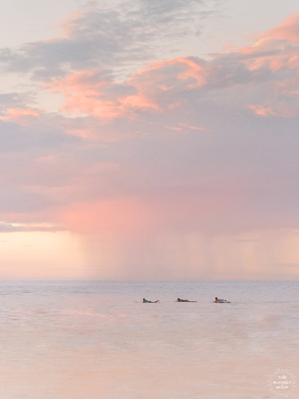 Surfers paddling in the ocean during a pastel pink rain squall at sunset in Costa Rica. What a surfer's dreams are made of - - beautiful sunset surf sessions. Have a piece of sunset surfer magic in your very own home with "Saltwater Paradise" sunset surfer wall art photographed by Kristen M. Brown of Samba to the Sea for The Sunset Shop.
